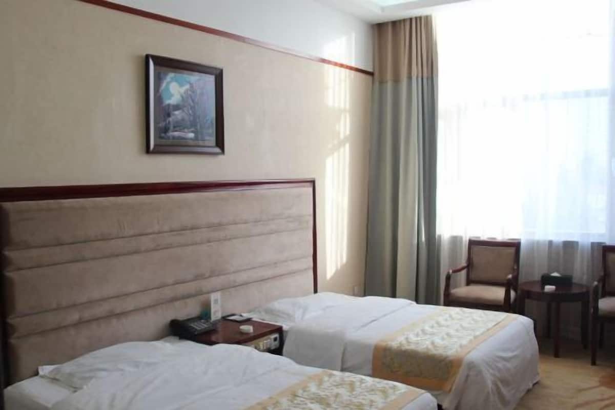 A hotel room with two beds, a nightstand, a framed picture on the wall, a window with curtains, and a seating area with chairs and a table.