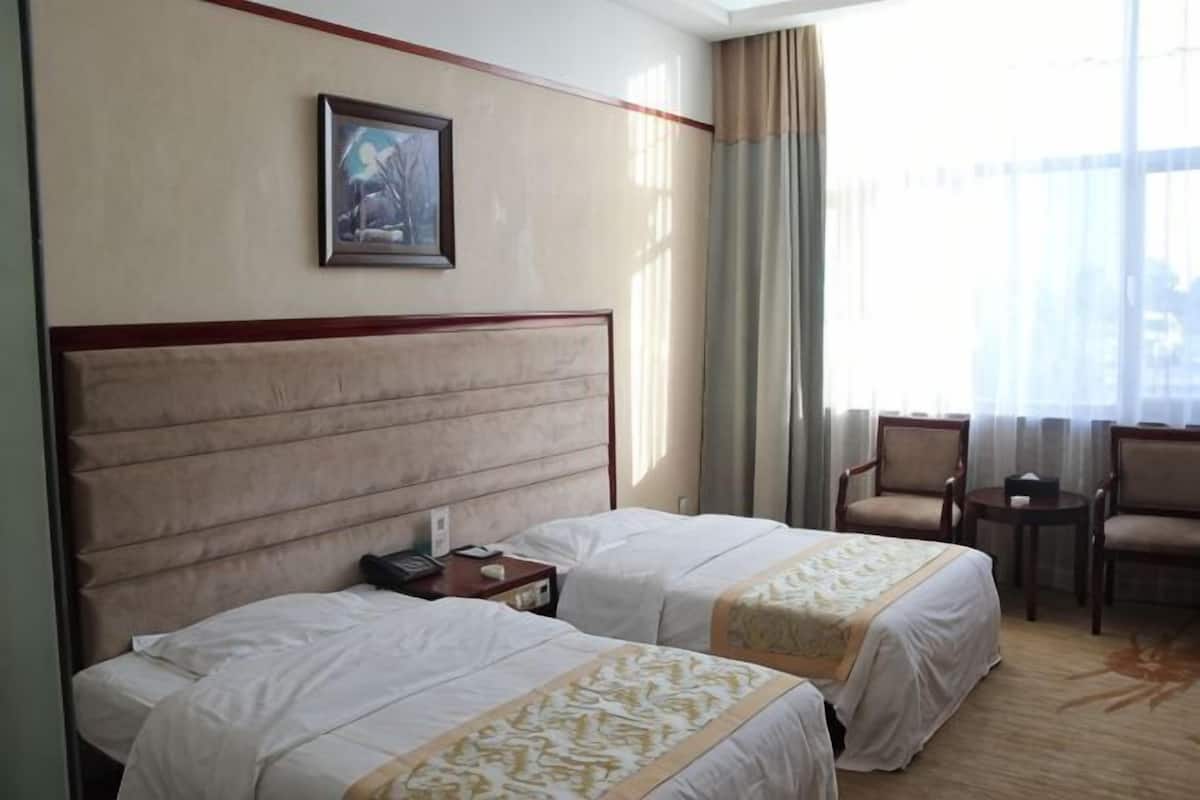 A hotel room with two beds, a small table, chairs, a painting on the wall, and a window with curtains.
