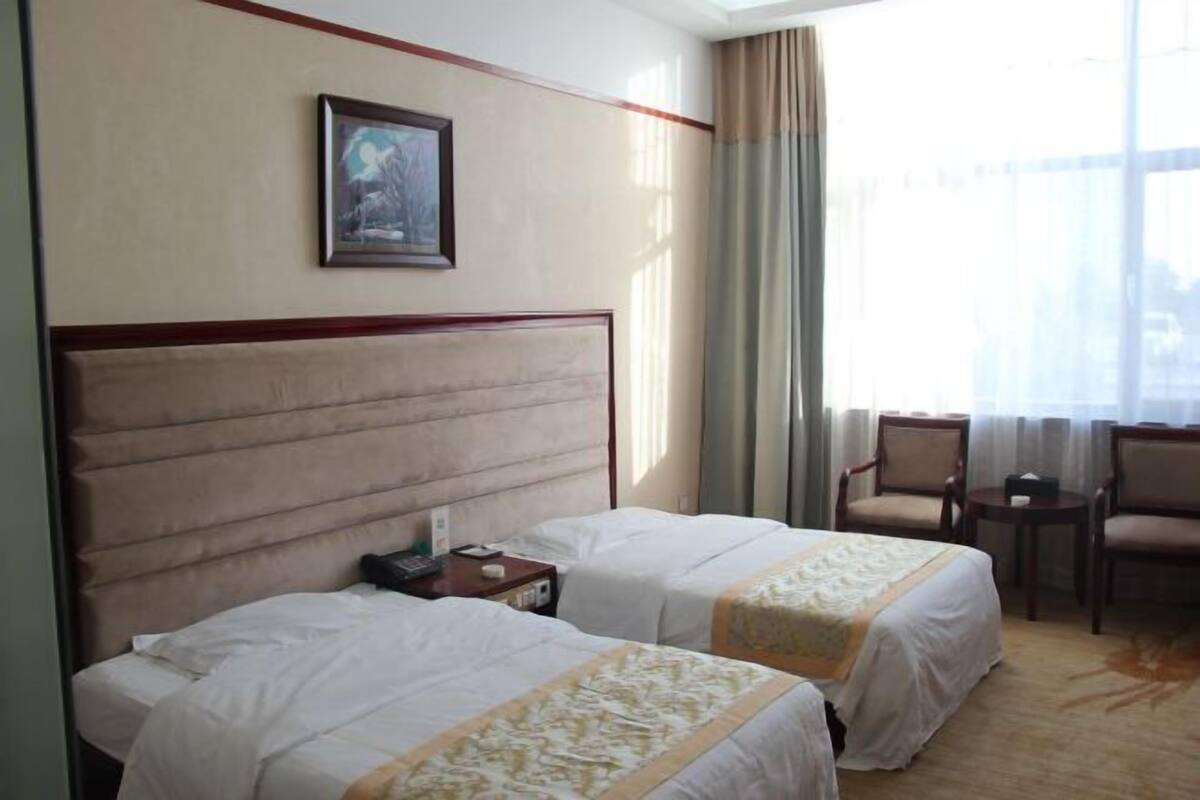 A hotel room with two beds, a painting on the wall, a window with curtains, and a small table with chairs.
