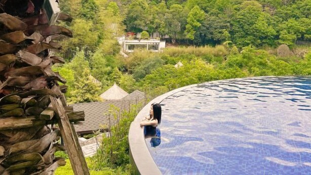 A pool with a curved edge, surrounded by lush greenery and a distant house. - Xiuhe Hot Spring Holiday Hotel (Xiushui)
