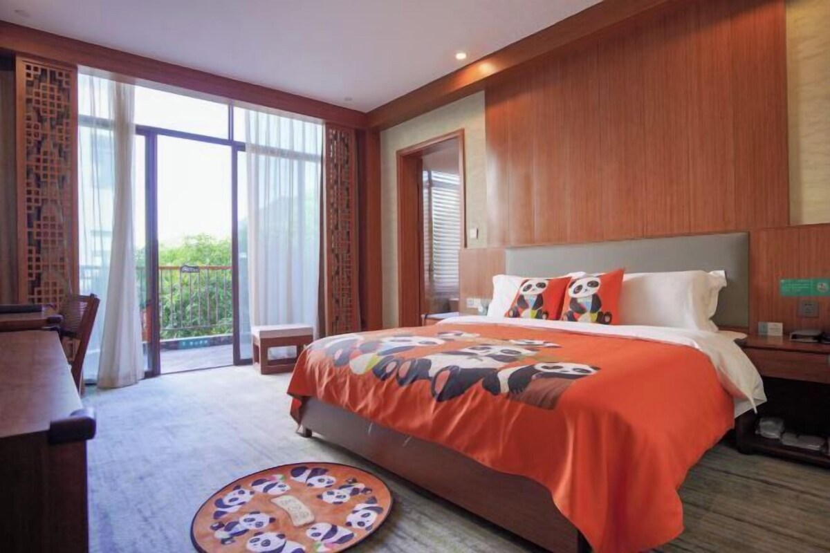 A hotel room with a large bed, a desk, and a view of greenery through the windows.