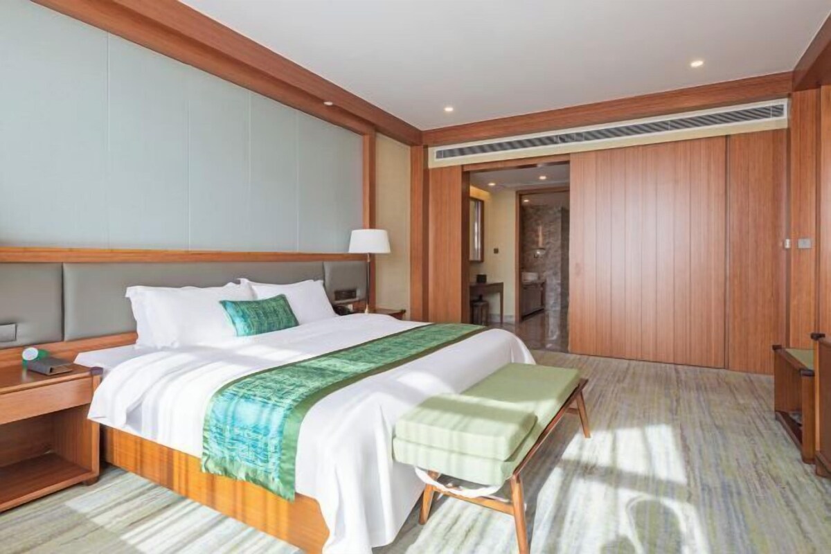 A hotel room with a large bed, wooden headboard, a bedside table, a bench, and a wardrobe.