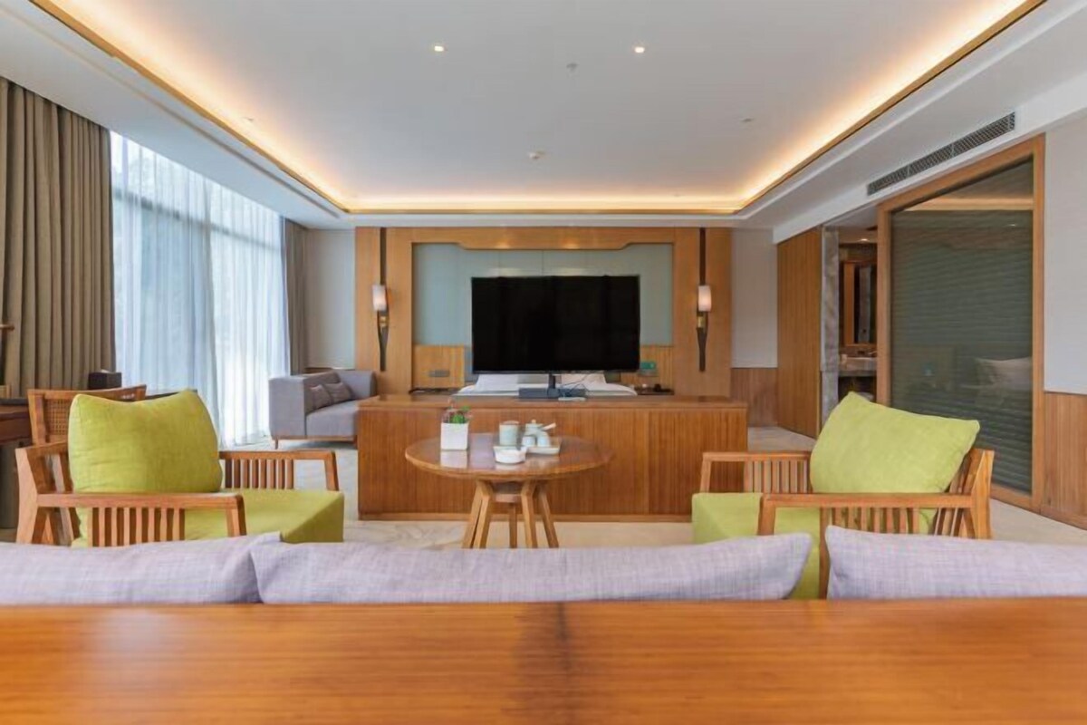 A modern hotel room with a wooden reception desk, a flat-screen TV, and a seating area with green cushioned chairs.