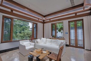 Amplio salón con sofá blanco, una mesa de centro de madera y grandes ventanas que dan a vistas de vegetación.