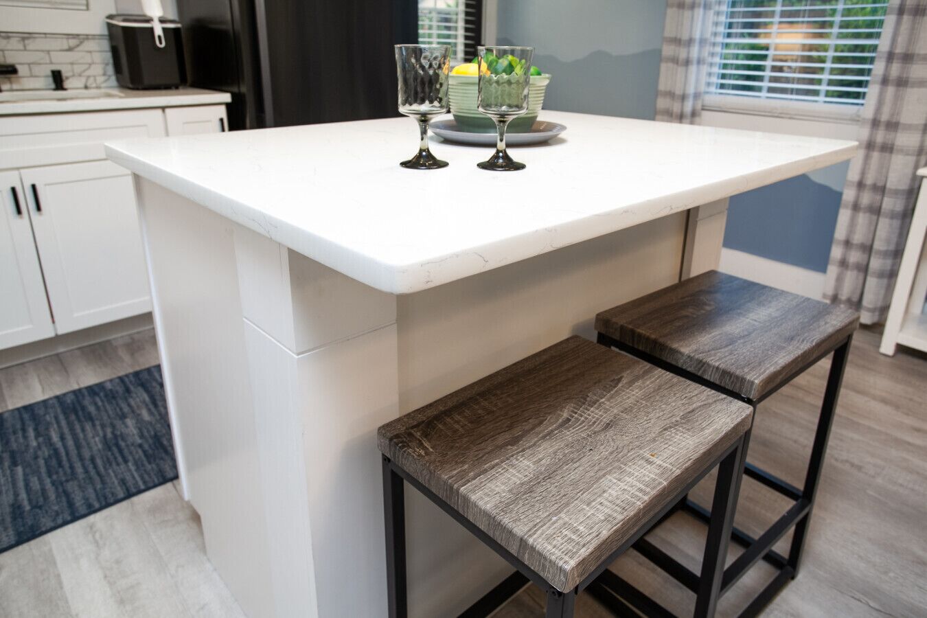 Una cocina con mostrador blanco, dos taburetes de madera y un recipiente de vidrio con limones y limas.