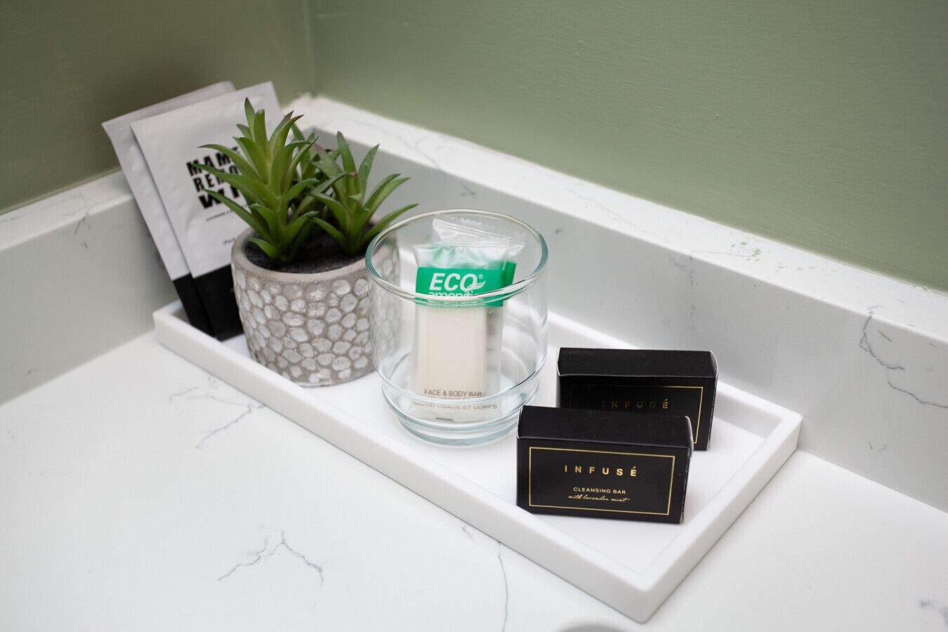 Un meuble-lavabo de salle de bain avec un plan de toilette en marbre blanc, un porte-bougie en verre portant la mention « ÉCO », une petite plante en pot et deux boîtes de savon noir.