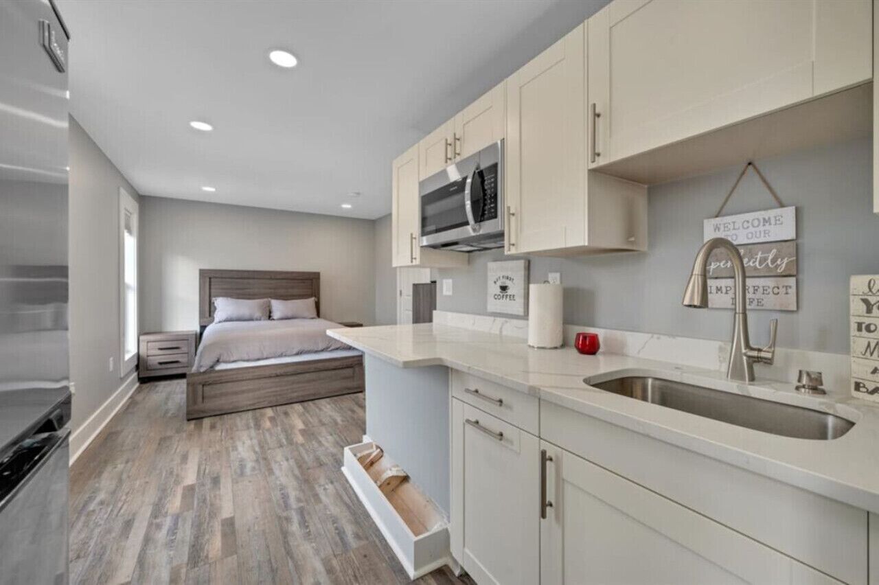 Un dormitorio compacto con una cama, una mesita de noche y un aparador. Junto al dormitorio hay una kitchenette con microondas, un lavabo y una encimera.