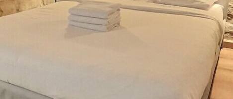 A neatly made bed with white linens and a folded towel, set in a room with stone walls and a wooden floor.