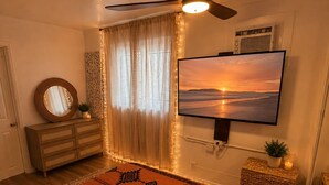 A bedroom with a bed, a ceiling fan, a TV, a dresser, a mirror, and a window with curtains. - The Zen Den (Venice)