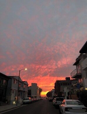 A street at sunset with buildings on either side and parked cars.