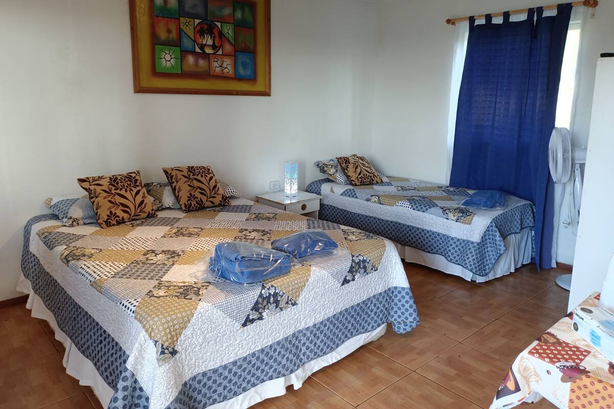 A room with two beds, a nightstand, a wall painting, a fan, and a table with a box.