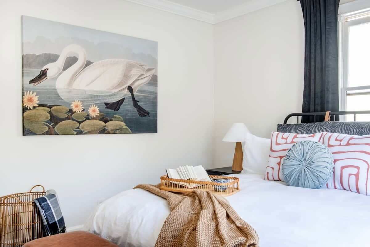 Un dormitorio con una cama, almohadas, una mesita de noche, una lámpara, una cesta y un cuadro de un cisne.