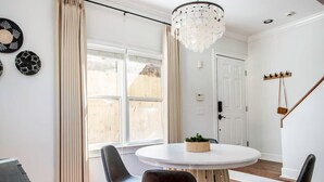 A dining area with a round table, two chairs, a chandelier, and a window with curtains. - Rook's Corner (Atlanta)