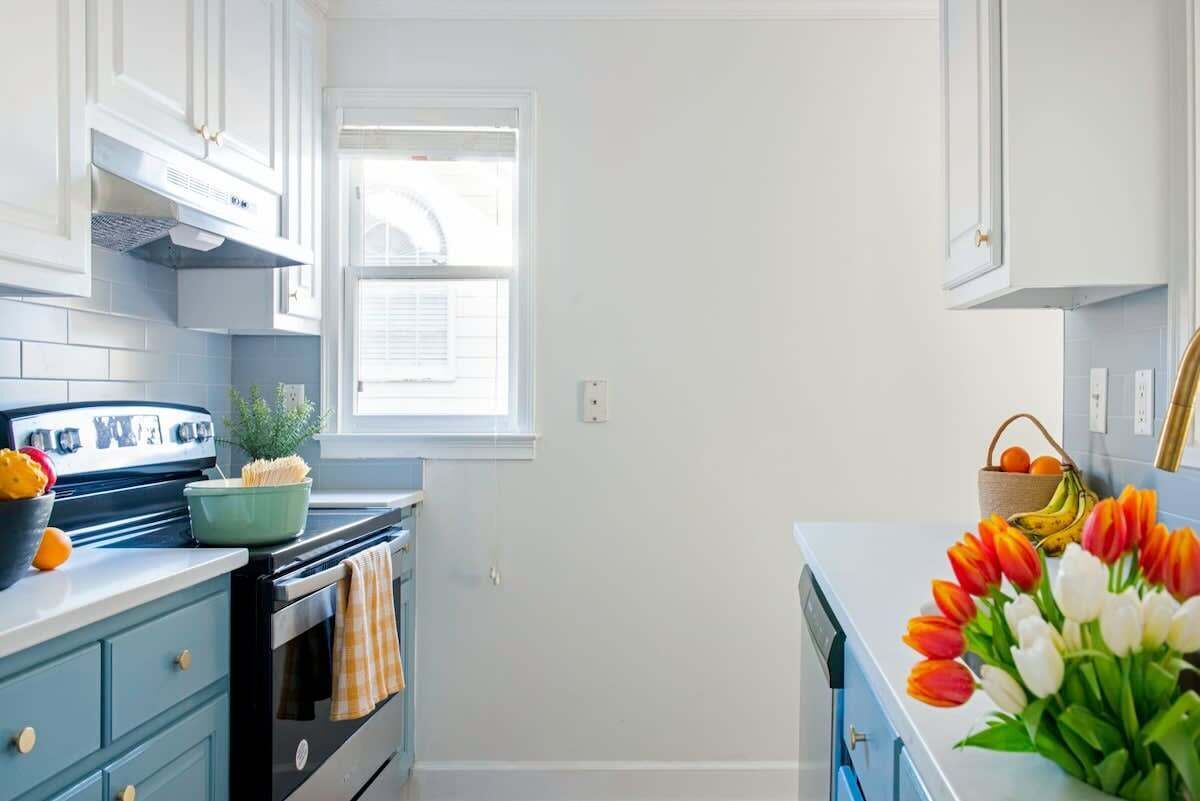 A kitchen with white cabinets, a stove, and a window. There is a bowl with fruits and a vase with flowers on the counter.