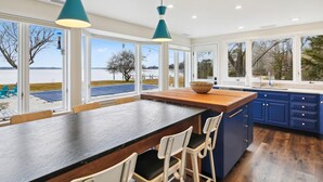 A modern kitchen with large windows, a wooden island, bar stools, and blue cabinets.