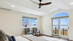 A bedroom with a large bed, two chairs, a ceiling fan, and a view of a lake through a glass door.