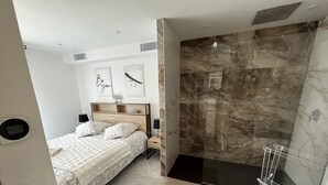 A modern bedroom with a bed, bedside tables, a nightstand, a shower area with a glass door, and a marble wall. - Charming Villa (Saint-Martin-du-Var)