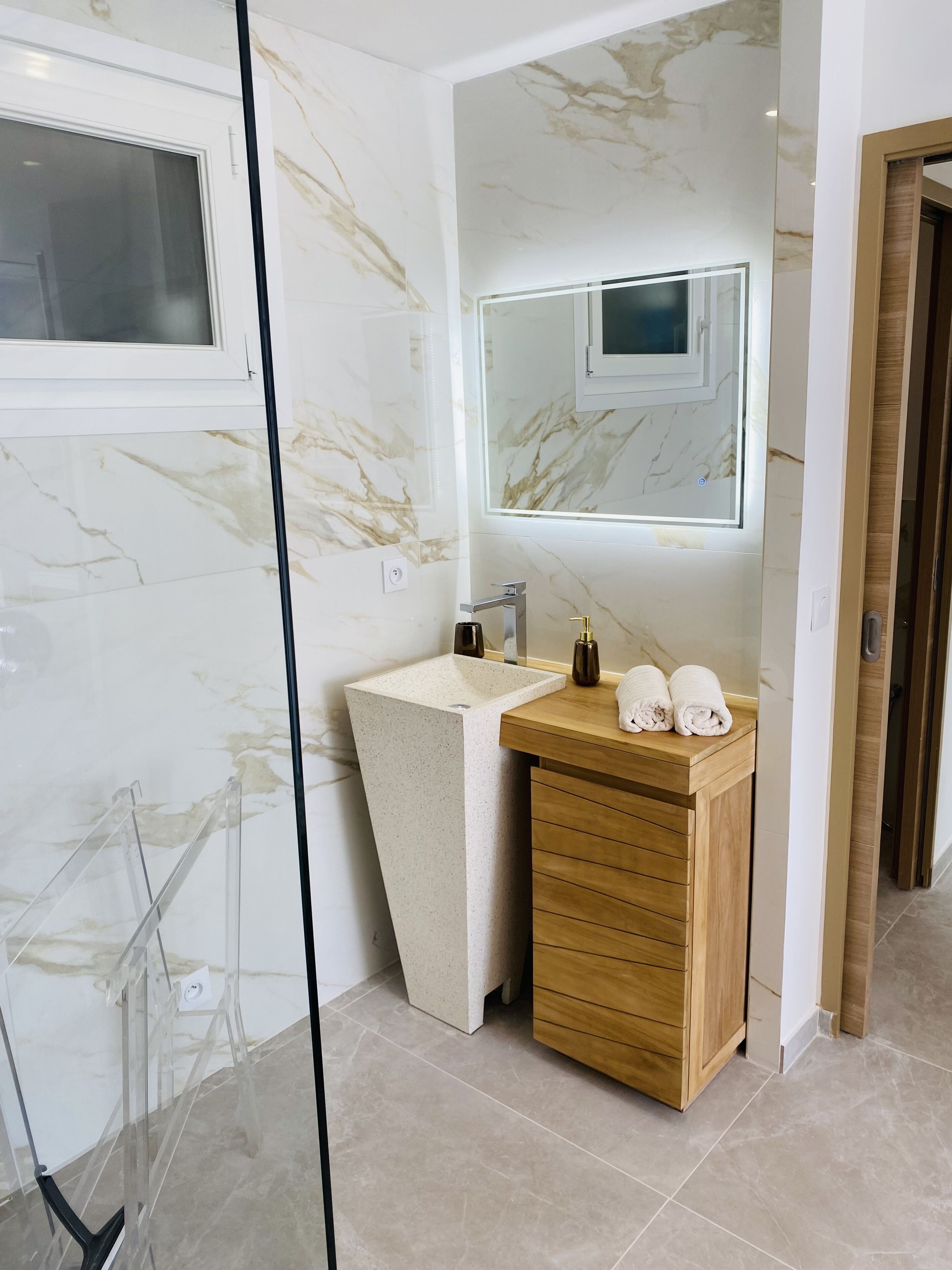 A modern bathroom with a marble backsplash, a wooden vanity, a large mirror, and a glass shower enclosure.