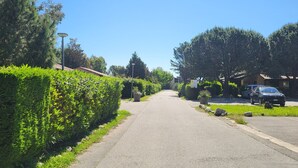 A residential street with a hedge-lined sidewalk, parked cars, and houses with sloped roofs. - Residence by the sea with private beach access, surrounded by greenery.  (Ghisonaccia)
