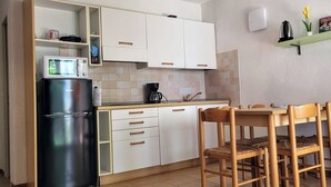 A compact kitchen with a refrigerator, microwave, and coffee maker. There are white cabinets with wooden handles, a tiled backsplash, and a wooden dining table with chairs. - Residence by the sea with private beach access, surrounded by greenery.  (Ghisonaccia)