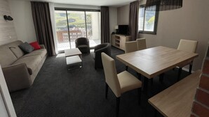 A modern hotel room with a sofa, dining table, chairs, a TV, and a large window offering a view of the outdoors. - 150m from ski slopes, 3rd floor, swimming pool, terrace, tv, ski locker, 49m², Gourette (Eaux-Bonnes)