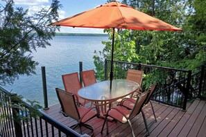 Een terras buiten met een ronde glazen tafel en vier stoelen, een rode parasol, en uitzicht op een meer.