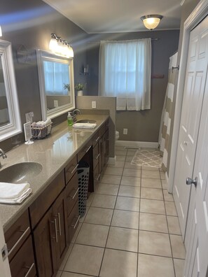 A modern bathroom with a vanity, sink, and mirror. It includes a towel rack, a trash bin, and a shower area with a curtain.