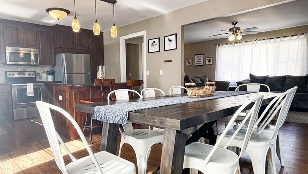A modern kitchen with stainless steel appliances, a dining area with white chairs and a wooden table, and a living room with a sofa and a ceiling fan. - 7th Heaven Guesthouse (Hermann)