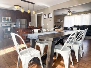 A modern kitchen with stainless steel appliances, a dining area with white chairs and a wooden table, and a living room with a sofa and a ceiling fan.