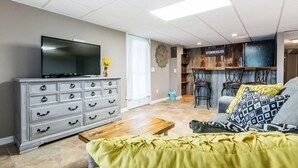 A living room with a TV, a wooden coffee table, a sofa with patterned cushions, and a kitchen area with bar stools. - 7th Heaven Guesthouse (Hermann)