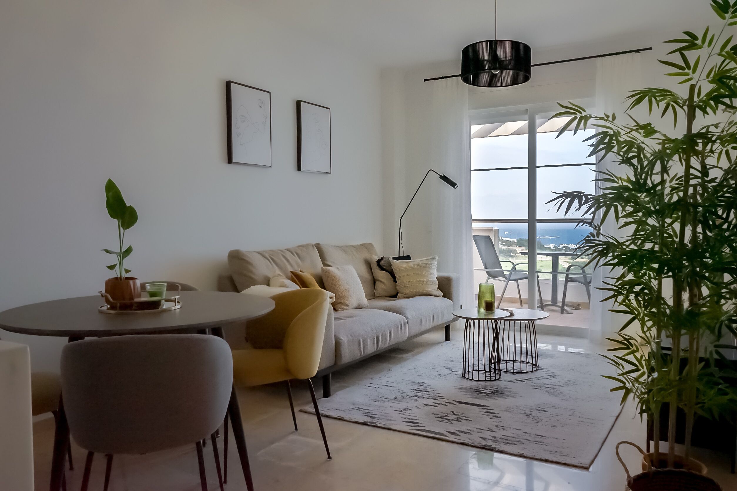 Un salon moderne avec un canapé, une table à manger et des chaises. Deux tableaux encadrés ornent le mur, ainsi qu’une lampe sur pied et une grande fenêtre offrant une belle vue.