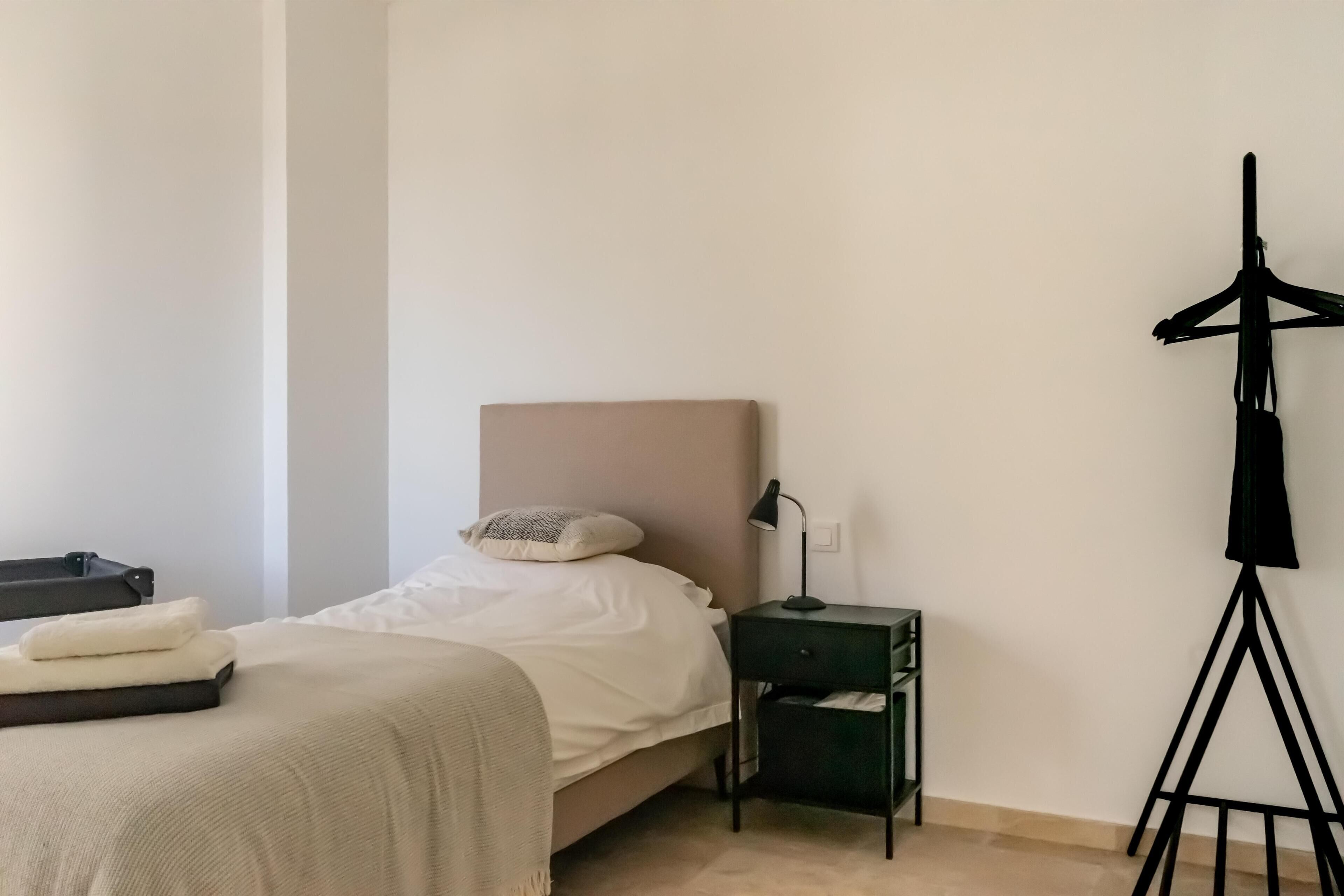 Een net opgemaakt bed met een hoofdbord, een nachtkastje met een lamp en een kledingrek.