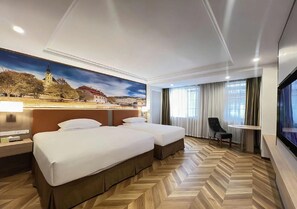 A hotel room with two beds, a desk, a chair, a TV, and a large window with curtains.