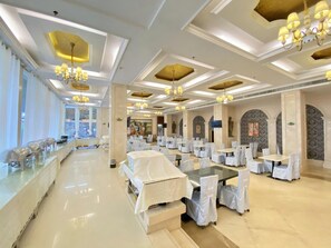 A spacious banquet hall with multiple tables and chairs, a long counter with glassware, and decorative lighting fixtures.