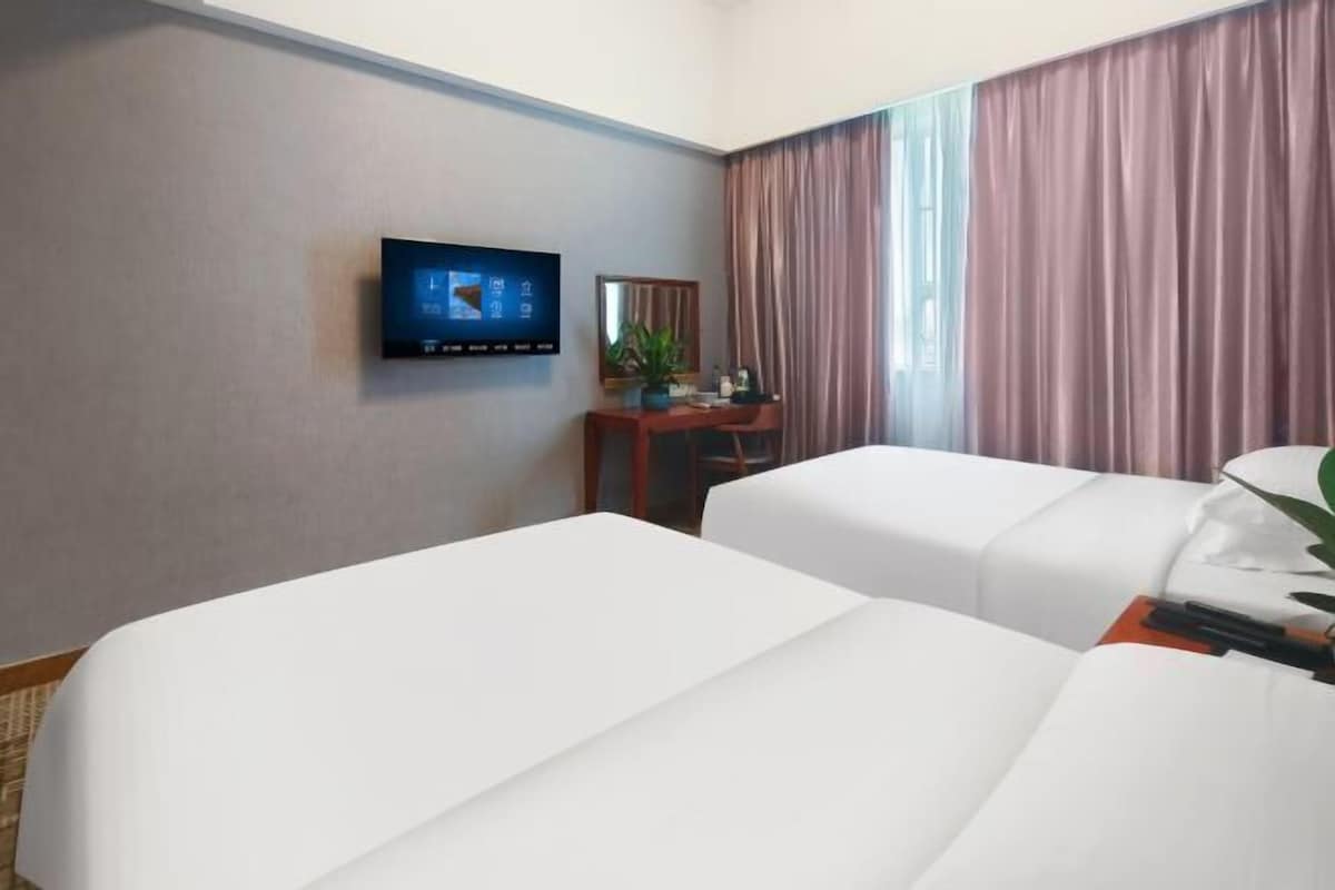 Quarto de hotel com duas camas, uma televisão na parede, uma mesa de trabalho com espelho e uma janela com cortinas.