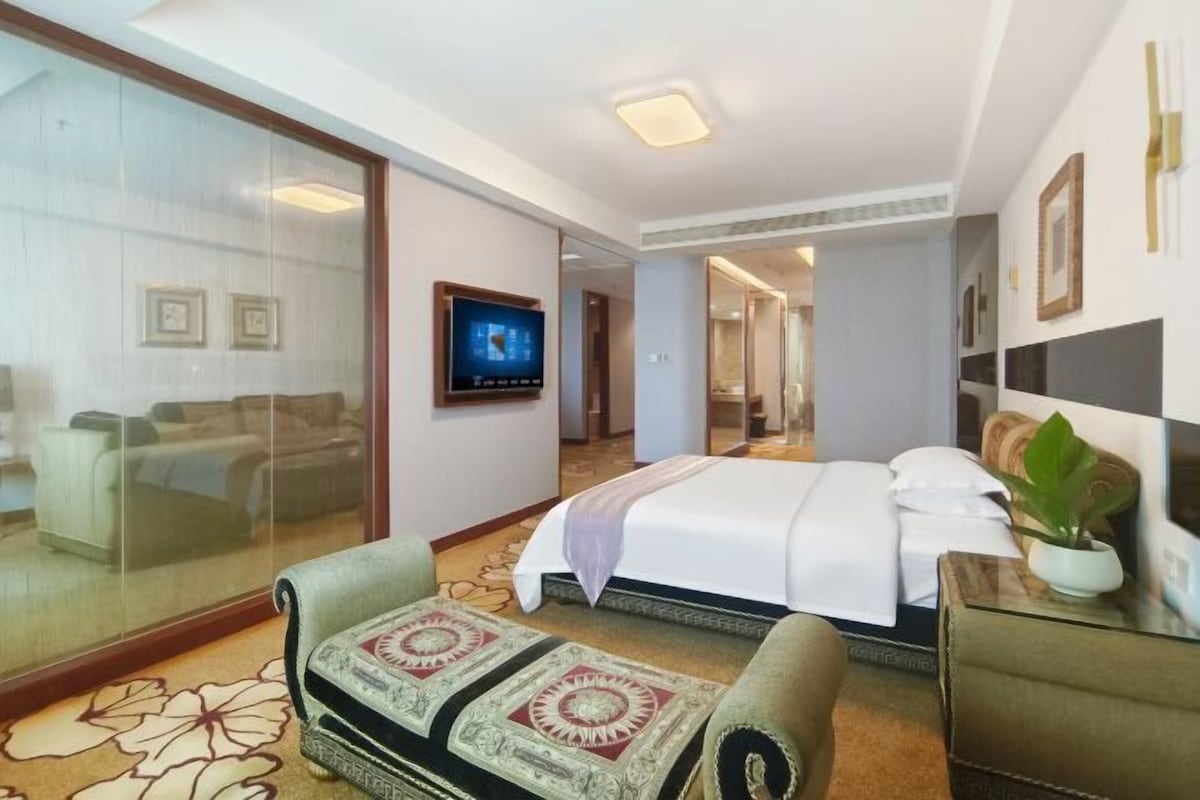 Quarto de hotel com cama, sofá, televisão e porta de vidro que dá para uma varanda.