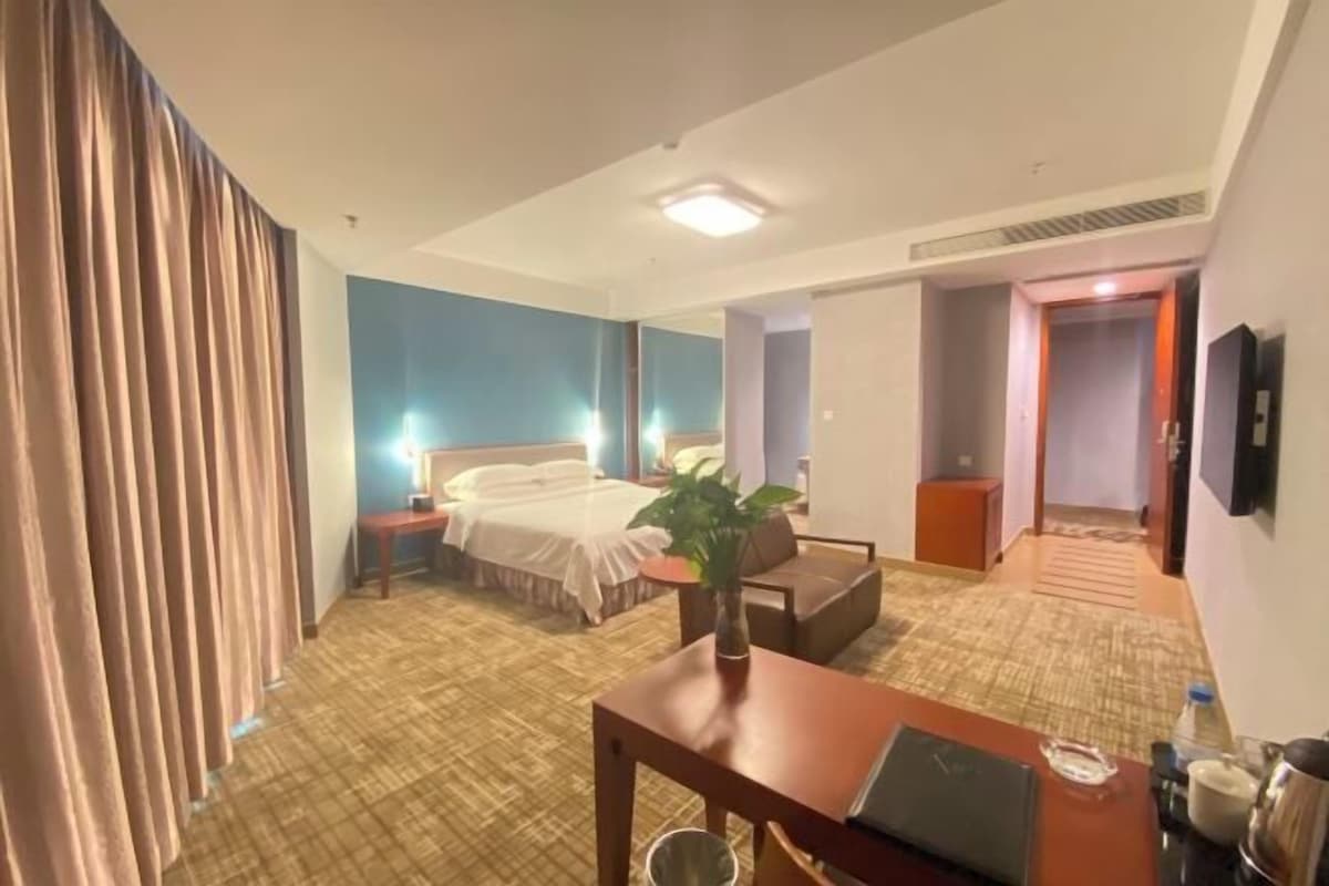 Quarto de hotel com cama, escrivaninha, cadeira, planta e banheiro visível por uma porta aberta.