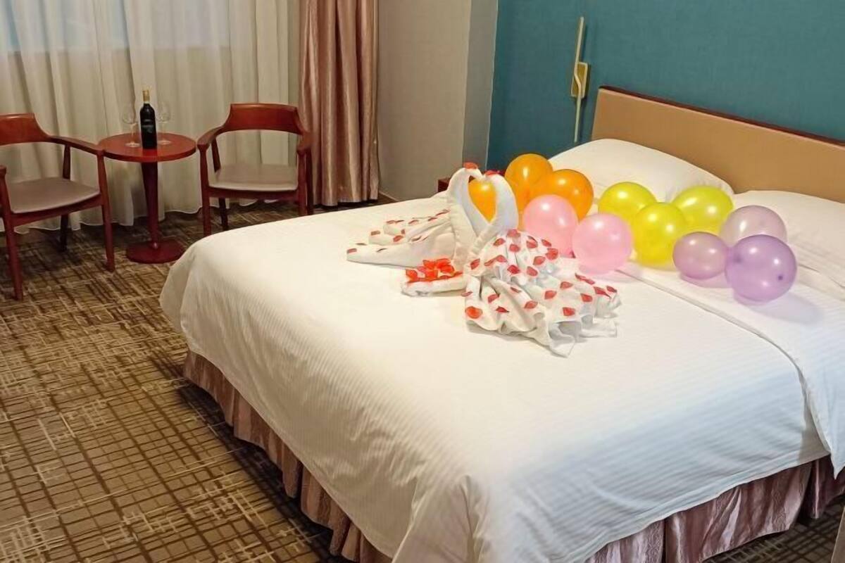 A hotel room with a bed, a small table with a bottle, two chairs, and balloons on the bed.