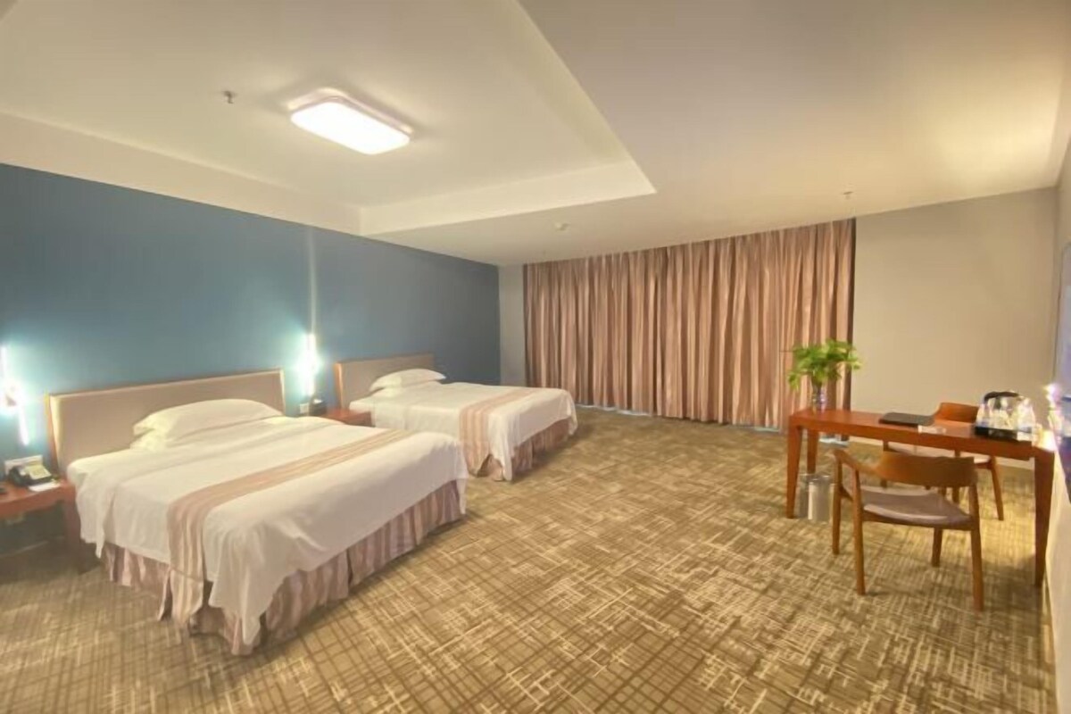 Quarto de hotel com duas camas, uma escrivaninha de madeira, uma cadeira e uma janela grande com cortinas.