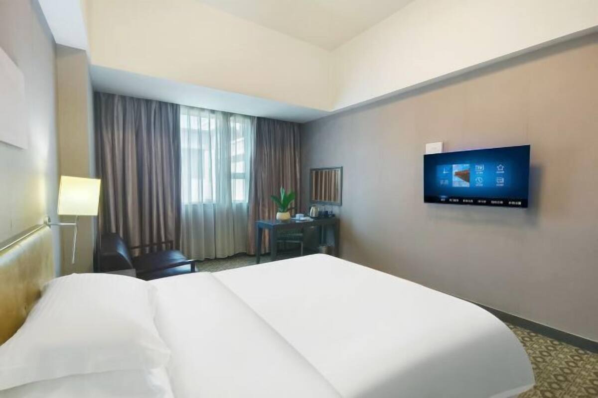 A hotel room with a bed, a desk, a chair, a TV mounted on the wall, and a window with curtains.