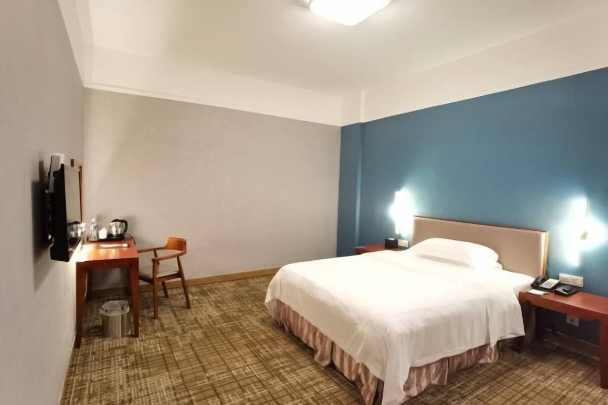 A hotel room with a bed, a desk with a chair, a trash bin, and a TV mounted on the wall.