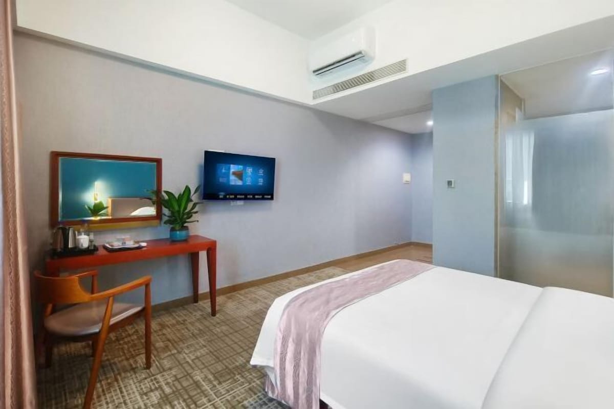 Quarto de hotel com uma cama, uma secretária com cadeira, uma televisão e um vaso de plantas.