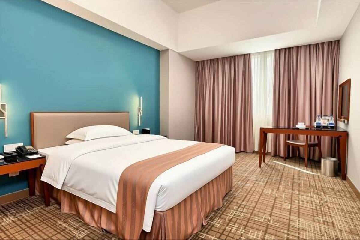 A hotel room with a bed, bedside table, desk, chair, and a large window with curtains.