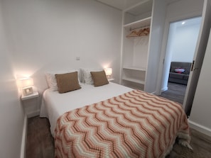 A neatly arranged bedroom with a bed, two bedside lamps, a wooden wardrobe, and a view of a living area.
