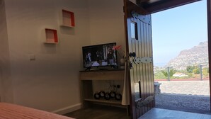 Ein Zimmer mit einem Bett, einem Fernseher auf einem Holzboden, einer zur Außenseite führenden Tür und einer kleinen Ablage mit Gegenständen.