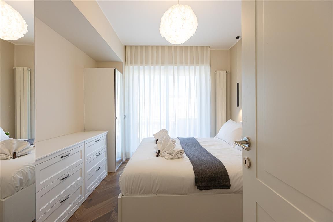 Una camera d'albergo con un letto, una cassettiera, uno specchio e un'ampia finestra con tende.