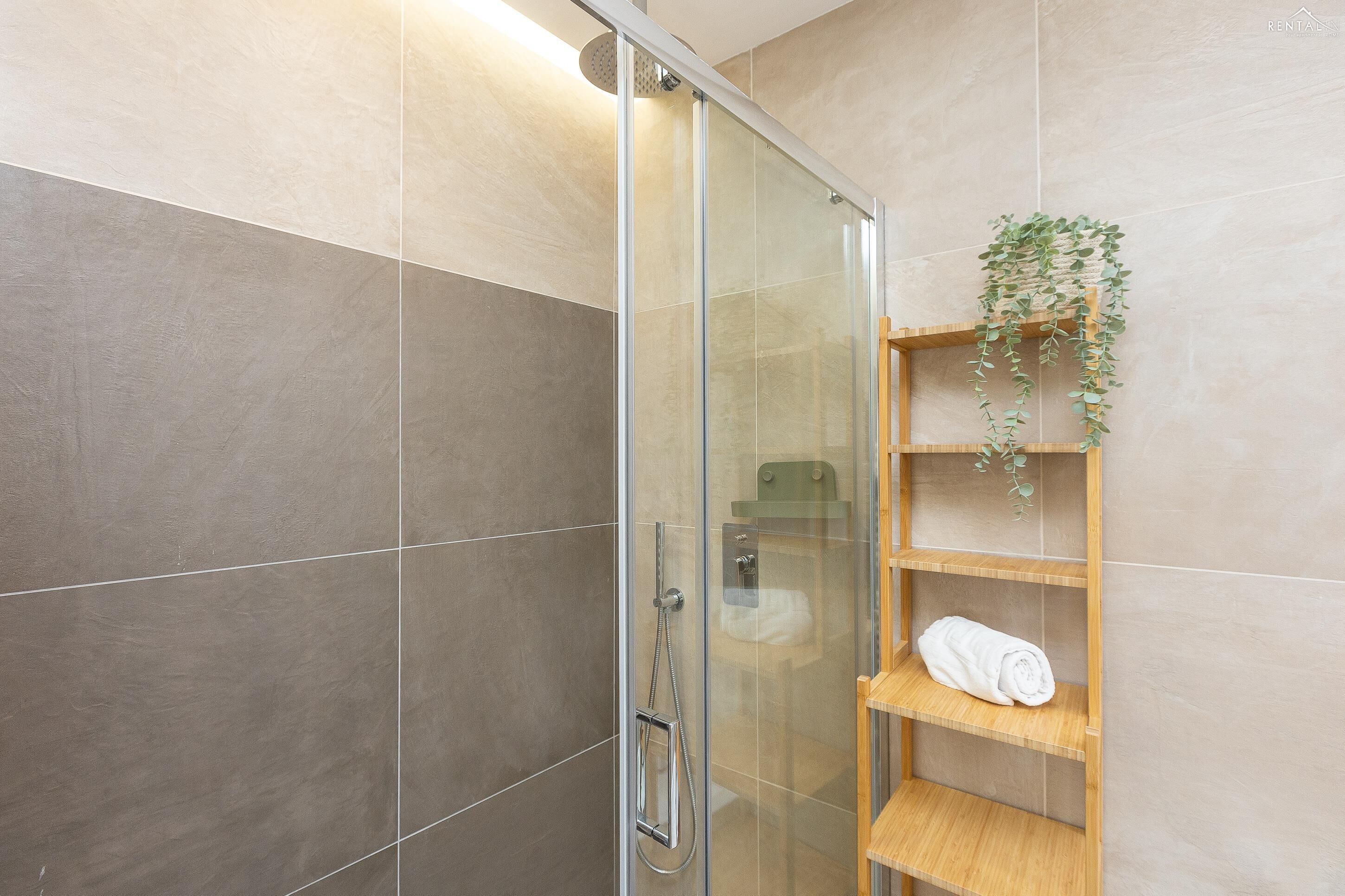 Un baño moderno con ducha envuelta en vidrio, un estante de madera con una toalla y una planta en maceta.