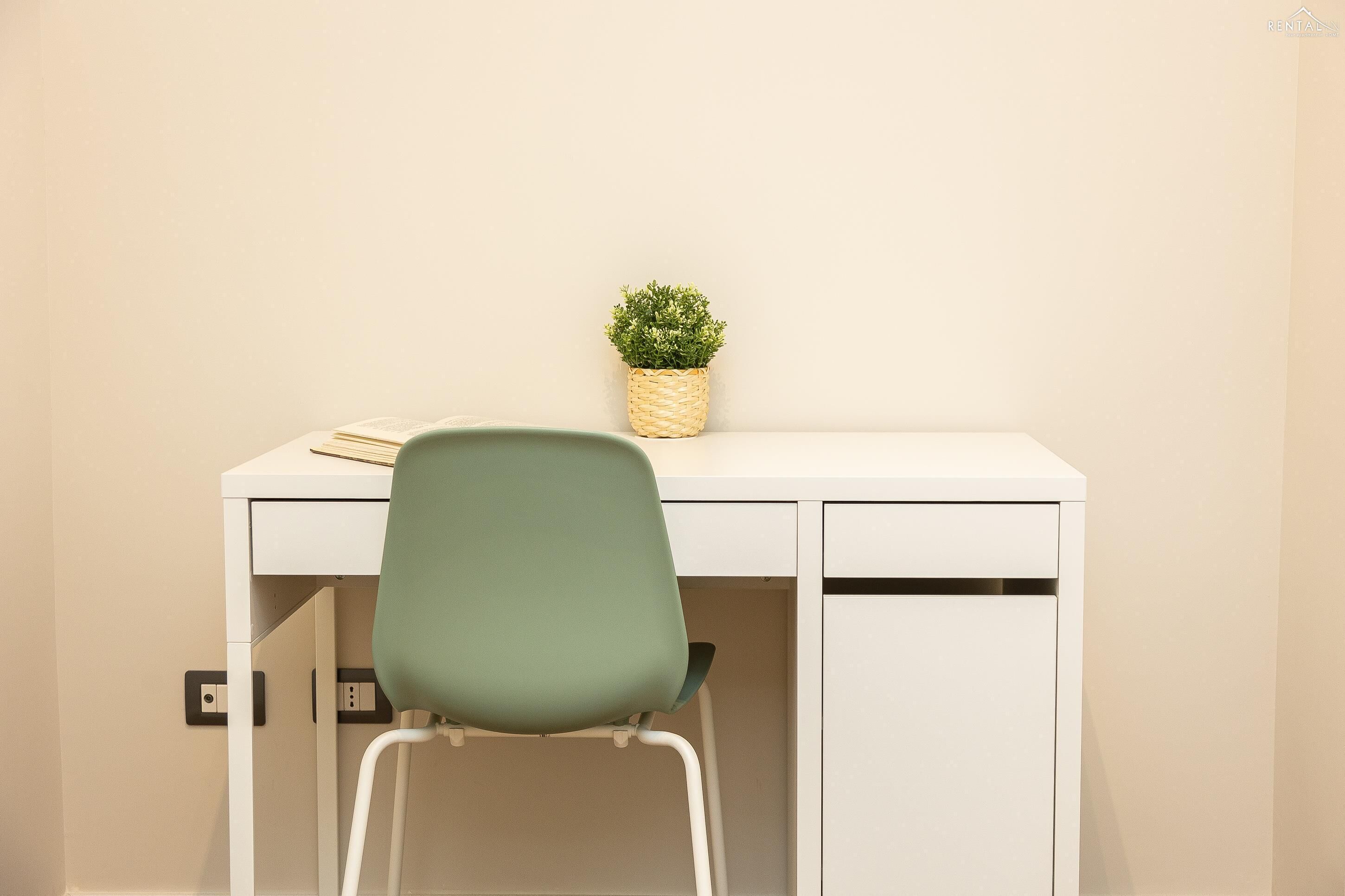 A white desk with a woven basket containing a small plant, a green chair, and a closed cabinet.