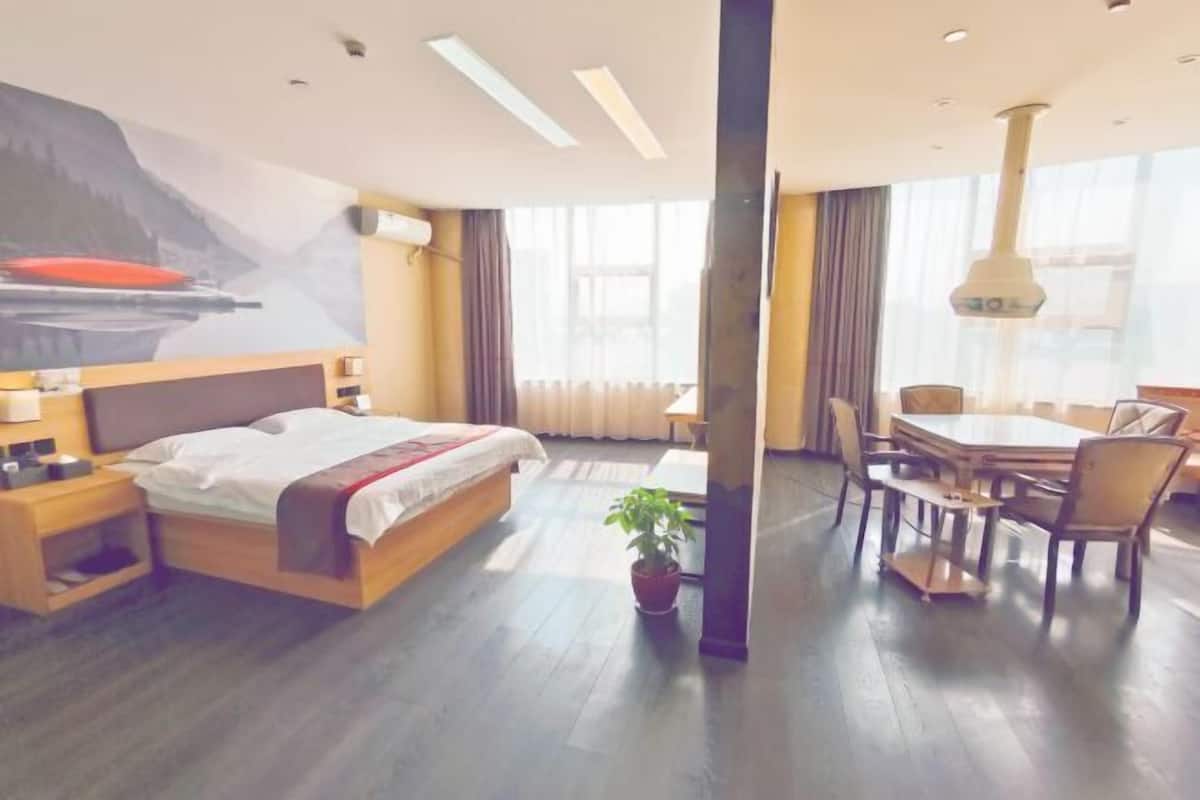 A hotel room with a bed, bedside table, lamp, potted plant, and a dining area with chairs and a table.