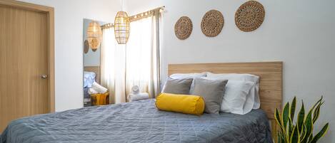 A neatly made bed with a grey quilt, a wooden headboard, and a yellow pillow. There are woven wall decorations and a potted plant to the side.